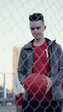 Vertical Screen: Portrait of a Handsome Young Football Player Looking at Camera, Spinning a Red Soccer Ball on His Finger. Stylish Footballer Standing Behind a Fence with a Group of Diverse People