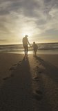 Vertical video of biracial father with son walking on sunny beach. healthy, active time beach holiday.
