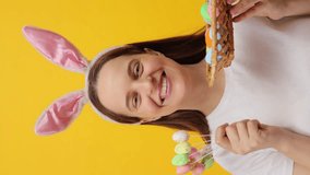 Vertical video. Beautiful happy young adult woman wearing bunny ears headband showing Easter eggs in basket and cake pops enjoying holiday celebration posing isolated over yellow background