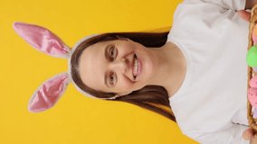 Vertical video. Easter-themed party for joyful moments. Celebrate the arrival of spring and Easter. Caucasian woman wearing rabbit ears headband posing isolated over yellow background