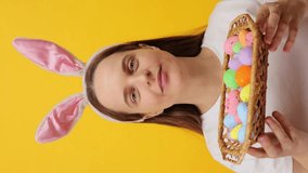 Vertical video. Good looking attractive young adult woman wearing bunny ears headband posing isolated over yellow background showing basket with Easter colored eggs