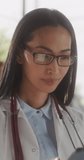 Vertical Screen: Portrait of a Beautiful Medical Health Care Professional Posing, Smiling and Looking at Camera. Asian Clinic Physician Wearing Glasses and a White Coat, Working in Hospital Office
