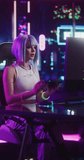 Vertical Screen: Young Woman in Futuristic Cosplay Clothes Sitting Down to Play Video Games on a Computer. Beautiful Girl Putting On Headphones and Talking to Players. Streaming and Gaming Concept