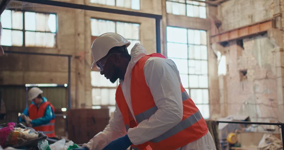 A man with Black skin in a special white protective uniform and an orange vest sorts plastic at an old waste processing plant. A man with Black skin is an employee of a huge waste recycling plant Royalty-Free Stock Footage #3453795071
