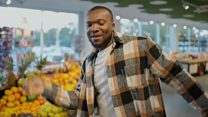 Portrait of a Happy man with Black skin color in a plaid shirt and a white T-shirt who is happy and dancing in a grocery store holding a grocery basket in his hands Royalty-Free Stock Footage #3453801875