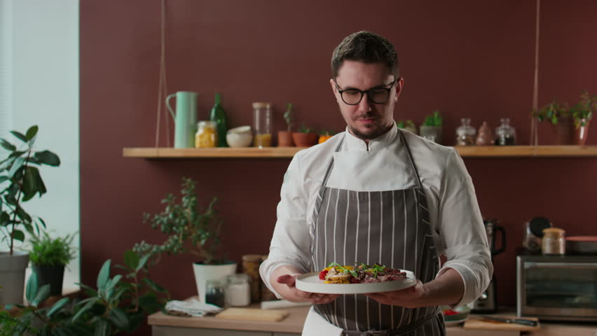 Medium portrait of Caucasian male chef wearing chefs uniform and eyeglasses posing for camera with delicious meat steak and grilled veggies on plate Royalty-Free Stock Footage #3453806449