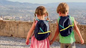 Two children with backpacks on Spain trip standing on a viewpoint of ancient Alhambra palace, pointing overlooking a panoramic view of a city and mountains in the distance - Powered by Shutterstock - Get 15% off with code: PIKWIZARD15