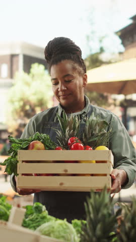Vertical Screen: Black Female Working at Farmers Market Stall with Fresh Organic Agricultural Products. African Businesswoman Holding a Crate with Fruits and Vegetables, Looking at Camera and Smiling Royalty-Free Stock Footage #3453845397