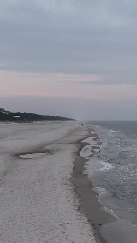 Coastline during pink sunset, gentle waves, white sand, looking at the horizon. Pastel colours of the sky, a  company of a pine forest and sand dunes with yellow coastal grasses. 