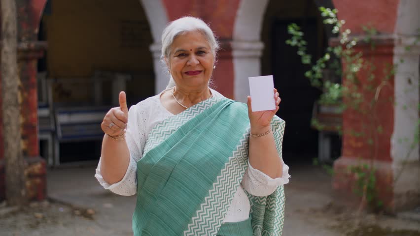 Grey-haired old lady dressed in saree, feeling happy after casting her vote in Indian elections - mature citizen. Smart old female showing her voter identification card while doing a gesture of thu...
