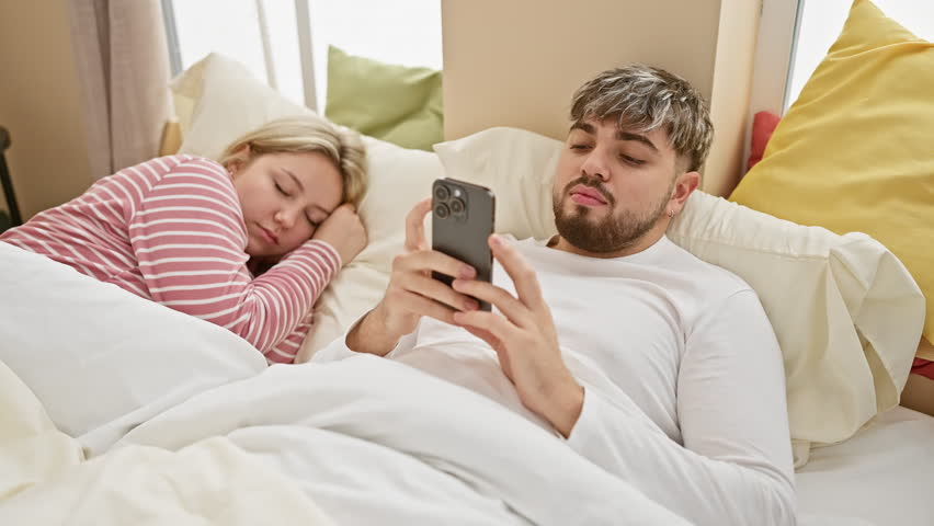 A man browsing his phone beside a sleeping woman in a cozy bedroom, depicting modern domestic life and relationships. Royalty-Free Stock Footage #3453882517