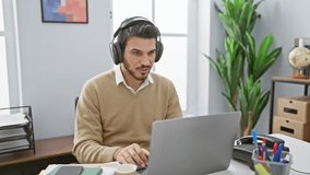 Hispanic man with beard wearing headphones celebrates success at computer in modern office.