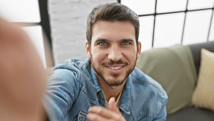 A handsome young hispanic man with a beard takes a selfie in a modern indoor living room.