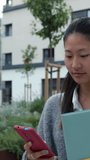 Vertical footage of happy young asian college student having fun using mobile phone at university campus. Millennial chinese female laughing while looking at smartphone screen walking in city street.