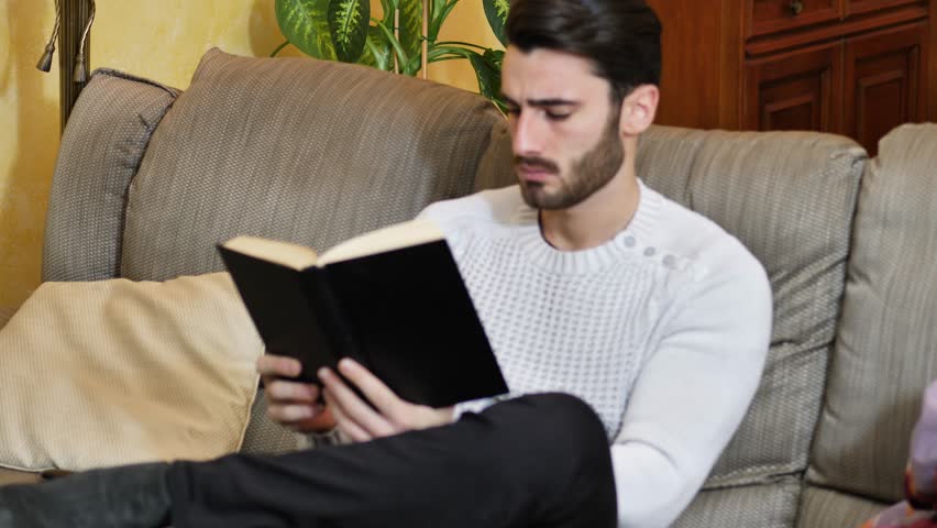Handsome black hair young man reading book at home, sitting on couch