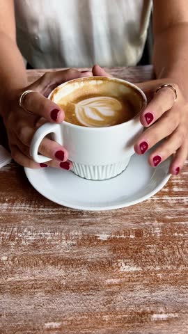 Casual coffee break details: close up of beautiful lady hands with red nails holding a white cup of coffee in a wood table