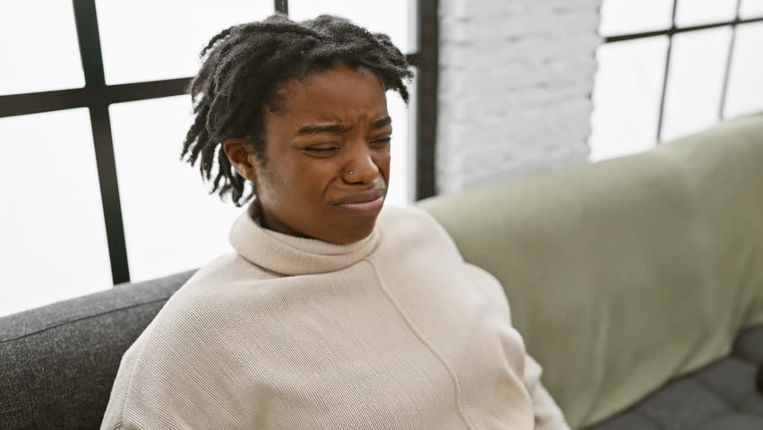 Young, skeptical black woman with dreadlocks frowning in discomfort on sofa at home, displaying a negative, problem-ridden expression. she's nervous, upset - an unhappy portrait of a doubtful doubter. Royalty-Free Stock Footage #3454060263