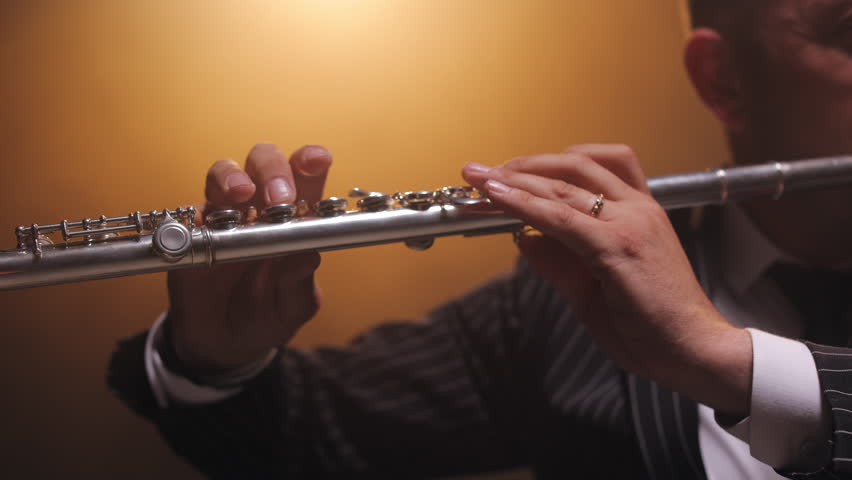 Crop of hands of skilled flutist holding instrument and creating stunning tune with soft finger movements. Attractive male in elegant suit playing music on flute in bright hall in front of audience.