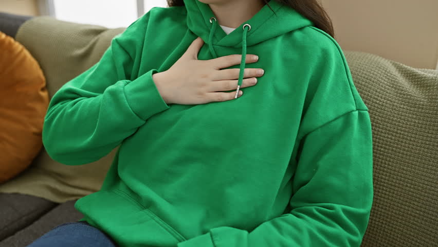A casual young woman in green hoodie relaxes at home on a comfortable couch, hand on chest, conveying a sense of wellbeing.