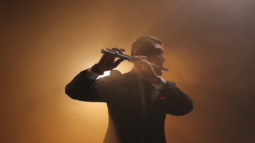 Low angle view of man holding musical instrument and performing while standing against bright spotlight. Creative male musician playing flute and expressing deep feelings through lyric music.