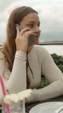 A young woman is talking on the phone sitting in a restaurant by the sea. Vertical video Social Media.