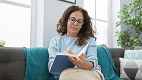 Mature hispanic woman with curly hair writing in notebook while sitting on couch indoors - Powered by Shutterstock - Get 15% off with code: PIKWIZARD15