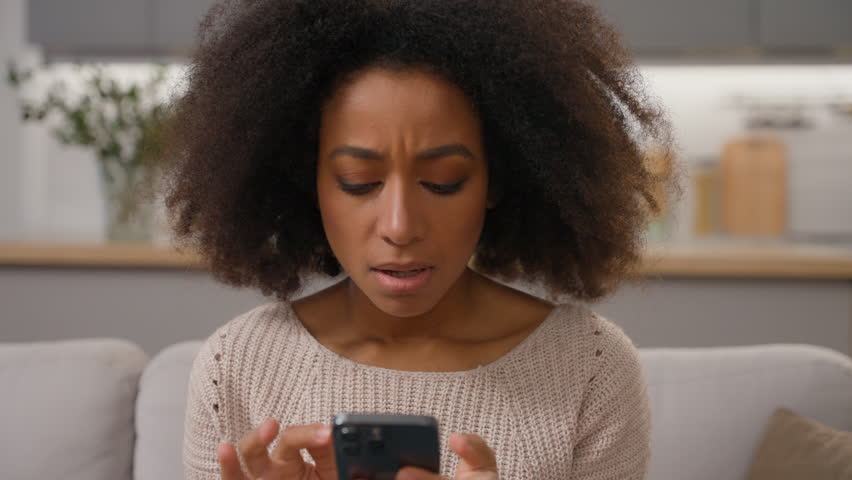 Worried frustrated African American ethnic woman on couch holding mobile phone in hands receive message bad news upset stressed anxious shocked girl female lady problem cell smartphone failure crash Royalty-Free Stock Footage #3454207401