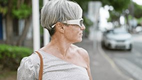 Elegant mature woman with grey hair and sunglasses walks confidently on a city street.