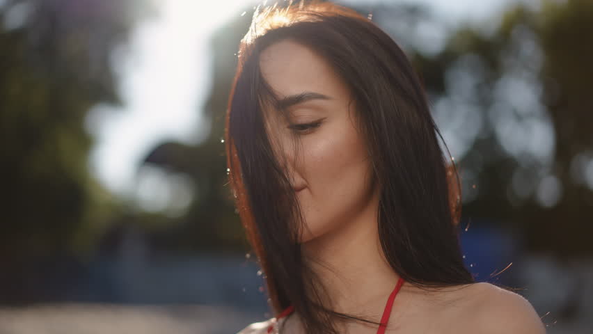 Slow motion. Close-up. The girl removes a strand of hair from face with her hand and smiles. A beautiful girl throws her head back, straightening her long hair with her hand, while standing outdoors