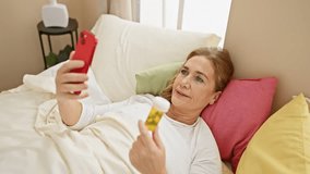 A middle-aged woman examines a pill bottle while video chatting on her smartphone in her bedroom