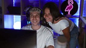 A woman leans over a man's shoulder in a neon-lit gaming room, sharing a joyful moment together indoors at night.
