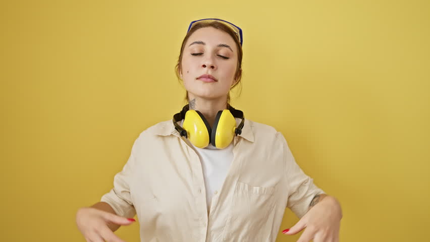Young brunette girl wearing safety glasses and noise reduction headphones relax and smiling with eyes closed doing meditation gesture with fingers. yoga concept. over isolated yellow background Royalty-Free Stock Footage #3454292649