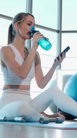 A woman is sitting on a yoga mat drinking water and looking at her phone