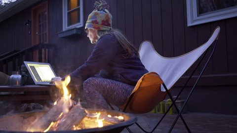 Woman sitting in butterfly chair working on computer outside in front of fire.