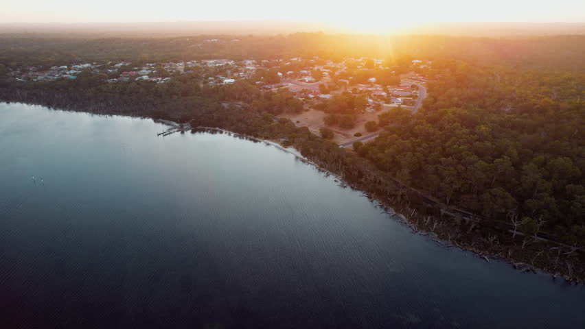Aerial view of a setting sun bathes a serene lakeside town in a warm golden light, quiet settlement nestles comfortably at the edge of the lake.