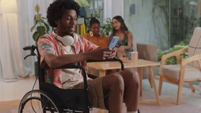 Portrait of young cheerful man in wheelchair using smartphone while gathering with friends at home, looking at camera - Powered by Shutterstock - Get 15% off with code: PIKWIZARD15