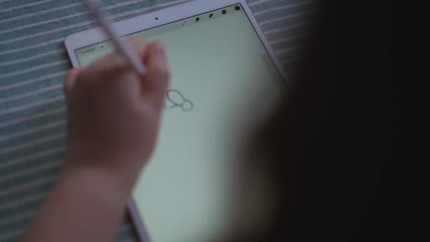View from shoulder of unrecognizable child sitting at a table in front of modern digital graphics tablet with stylus in hand, drawing using technological device. Shooting in slow motion.
