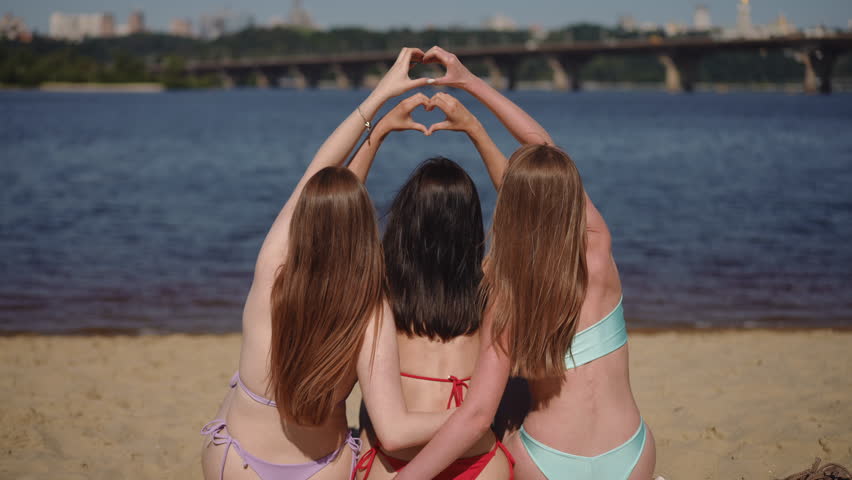 View from the back. Three girls in swimsuits sit on the sandy beach, hugging each other and raising their hands to make two hearts shape