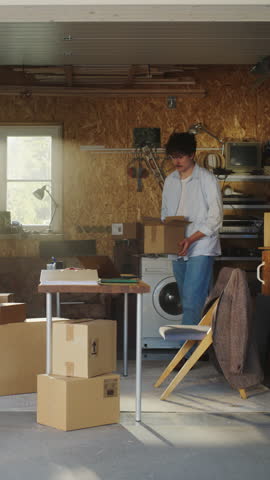 Vertical Screen: Caucasian Businessman Using Laptop In Garage. Small Business Owner Filling Orders From Online Clients. Packing Products In Cardboard Boxes For Shipping. Entrepreneur Working At Home.