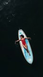 Aerial drone view on well looking middle aged woman with black hair in red swimsuit, swimming on sup in calm azure sea. Summer holiday vacation and travel concept. Vertical video
