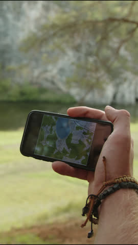 Vertical close up shot of hands of hiker looking at map on smartphone and pointing at direction while traveling in nature