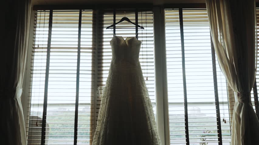 A wedding dress hangs on a wooden hanger by the window