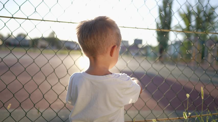boy stands near the playground. concept of happy childhood and loving family. a child holds on to the net of a sports ground, court and glare of the sun in the lifestyle background