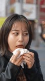 Vertical video of a young Japanese Okinawan woman in her 20s eating Okinawan traditional sweets lots of sata andagi on Kokusai Street, a tourist attraction in Naha City, Okinawa Prefecture