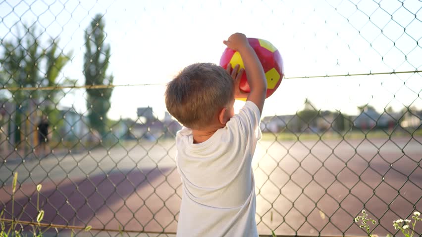 the boy stands near the playground. concept of happy childhood and loving family. child holding a red ball in his hands, playground and glare of the sun lifestyle in the background Royalty-Free Stock Footage #3454589919