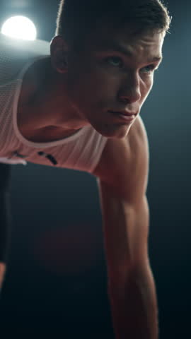 Vertical Screen: Strong Young Man Starting a Race From Track Starting Blocks Position on a Dark Stadium in the Evening. Cinematic Portrait of a Fit Male Sprint Runner Participating in a Competition