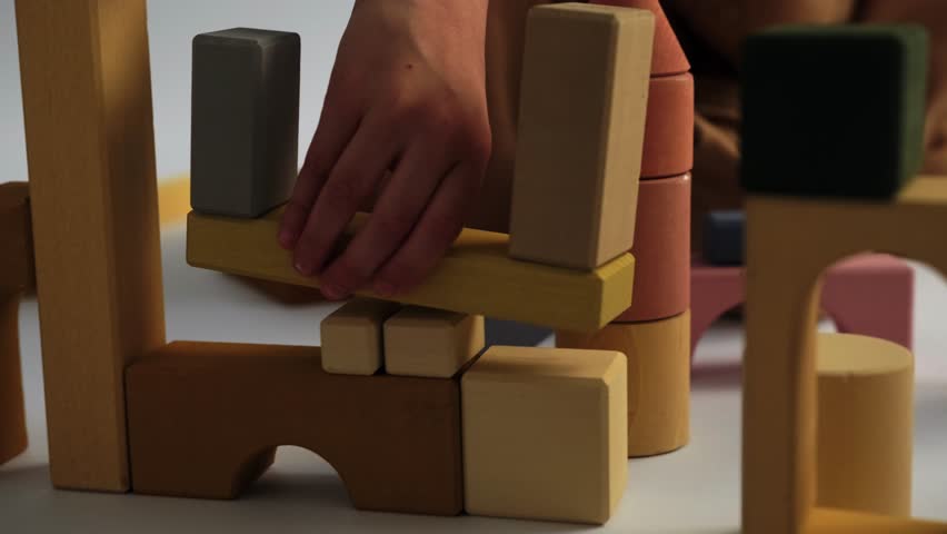 A blond boy sits on a white background and plays with a constructor, building a city from wooden blocks with multi-colored towers. Wooden construction set, childhood, educational games.