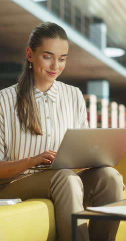 Vertical Screen: Smart College Student Using Laptop Computer to Study in a Public Library. Young Woman Learning Online, Getting Ready for Exams, Drafting an Essay While Sitting on a Sofa Royalty-Free Stock Footage #3454610223