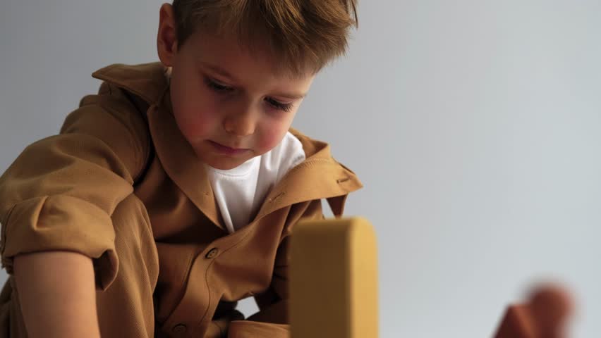 A blond boy sits on a white background and plays with a constructor, building a city from wooden blocks with multi-colored towers. Wooden construction set, childhood, educational games.