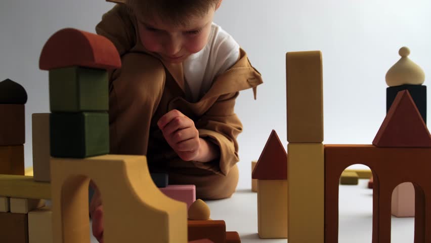 A blond boy sits on a white background and plays with a constructor, building a city from wooden blocks with multi-colored towers. Wooden construction set, childhood, educational games.